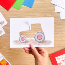 Load image into Gallery viewer, A hand holds a printed card featuring a watercolor-style tractor cut-out with gray wheels and red-rimmed tires, set on a wooden table with craft supplies around it.
