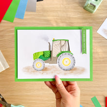 Load image into Gallery viewer, A hand holds an iris fold greeting card featuring a green tractor illustration with a white background and green border, set on a wooden desk with craft supplies around it.
