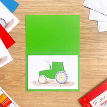 Load image into Gallery viewer, A green greeting card featuring a watercolor cut-out tractor on a white panel, displayed on a light wood tabletop with colorful envelopes and stationery props around it.
