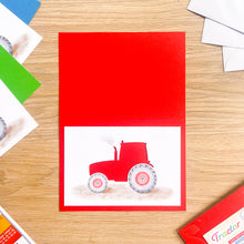 Load image into Gallery viewer, A red greeting card featuring a watercolor-style illustration of a red tractor on a white background, placed on a light wood tabletop with other cards and envelopes partially visible around it.
