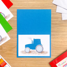 Load image into Gallery viewer, A handmade blue greeting card featuring a watercolor illustration of a blue tractor cut-out on a white background, displayed on a light wood tabletop with craft supplies.
