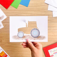 Load image into Gallery viewer, A hand holds a printed card featuring a watercolor-style tractor cut-out on a white background, set on a wooden tabletop with craft supplies around it.
