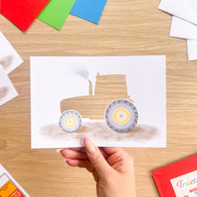 Load image into Gallery viewer, A hand holds a printed card aperture cut-out featuring a watercolor-style tractor illustration on a white background, photographed over a wooden tabletop with craft paper and colored cards around it.
