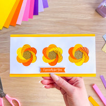 Load image into Gallery viewer, Handmade card with floral iris fold design held by a hand on a wooden surface with craft materials.
