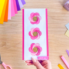 Load image into Gallery viewer, Hand holding a card with pink iris fold flowers on a wooden surface with craft supplies.
